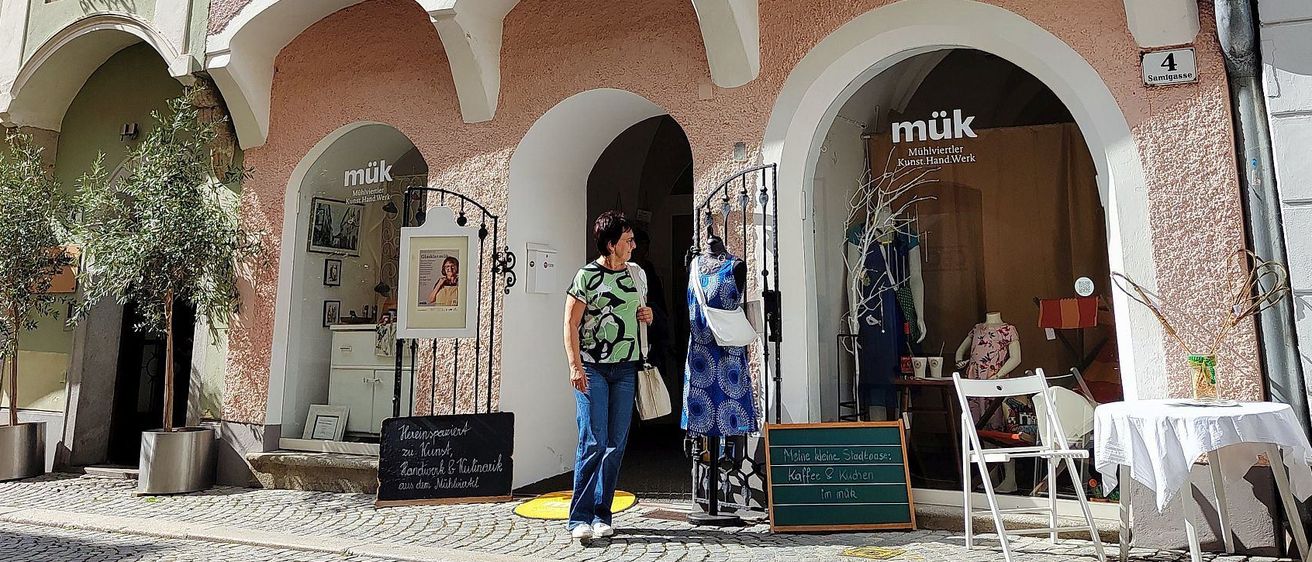 A woman stands in front of a boutique called muk. The boutique has glass doors and windows with white arches. There are two blackboards with writing and a potted plant outside.