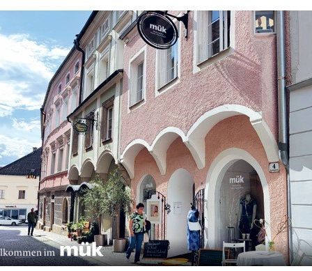 Eine Straßenszene mit einem Geschäft namens 'muk', einer Frau, die vorbeigeht, einem geparkten Auto und einer Person, die draußen steht.