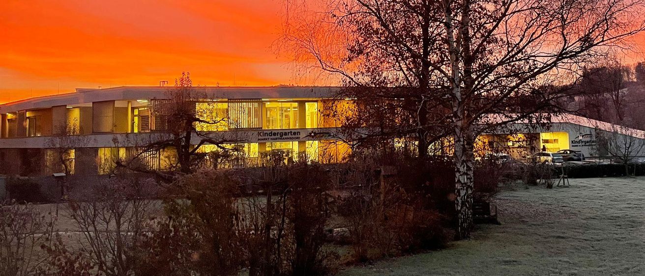 Ein Kindergartengebäude mit dem Schild 'Kindergarten Mark Hartmannsdoerf' ist bei Sonnenuntergang beleuchtet. Das Gebäude ist von Bäumen und Büschen umgeben. Der Himmel ist orange.