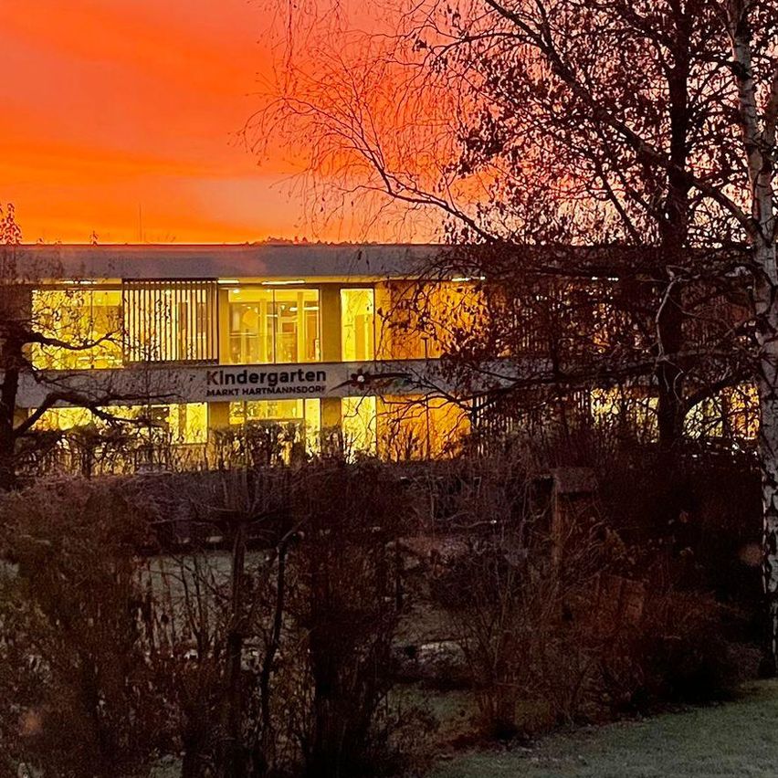 Ein Kindergartengebäude mit dem Schild 'Kindergarten Mark Hartmannsdoerf' ist bei Sonnenuntergang beleuchtet. Das Gebäude ist von Bäumen und Büschen umgeben. Der Himmel ist orange.