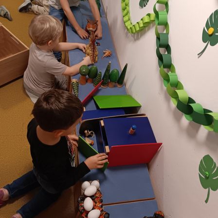 Zwei Jungen spielen mit Spielzeug auf einem blauen Regal. Einer sitzt und der andere steht. Auf dem Regal befinden sich Dinosaurierspielzeug, ein Baum und eine Dinosaurierfigur. An der Wand befindet sich eine Kette aus grünen Papierkreisen.