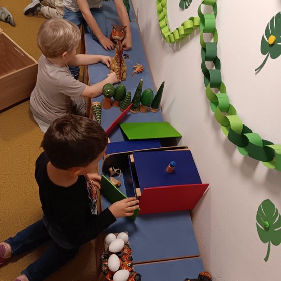 Zwei Jungen spielen mit Spielzeug auf einem blauen Regal. Einer sitzt und der andere steht. Auf dem Regal befinden sich Dinosaurierspielzeug, ein Baum und eine Dinosaurierfigur. An der Wand befindet sich eine Kette aus grünen Papierkreisen.