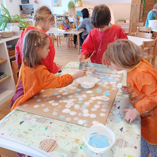 Fünf Kinder malen auf einer großen Leinwand mit blauen Kreisen. Sie tragen orangefarbene Lätzchen und sitzen um einen Tisch herum. Der Raum hat Holzböden und weiße Wände.
