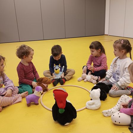 Mehrere Kinder sitzen in einem Kreis um einen Hula-Hoop mit Spielzeug auf dem Boden, darunter ein Hund, ein Bär und eine Katze.