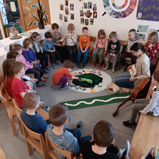 Eine Gruppe junger Kinder sitzt im Kreis um eine Frau mit einer Gitarre und spielt mit einem grünen Teppich auf dem Boden.