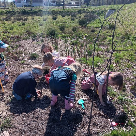 Kinder arbeiten in einem Grasbereich im Freien, eines hält einen roten Eimer. Sie tragen Stiefel und Jacken. Ein Baumstamm ist in der Nähe.