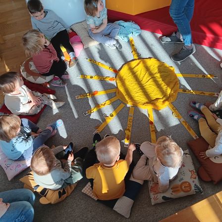Eine Gruppe von Kindern sitzt um eine gelbe Sonne auf dem Teppich herum und macht eine Aktivität. Sie tragen Freizeitkleidung, und einige tragen Turnschuhe. Eine Person in Jeans steht in der Nähe der Wand.