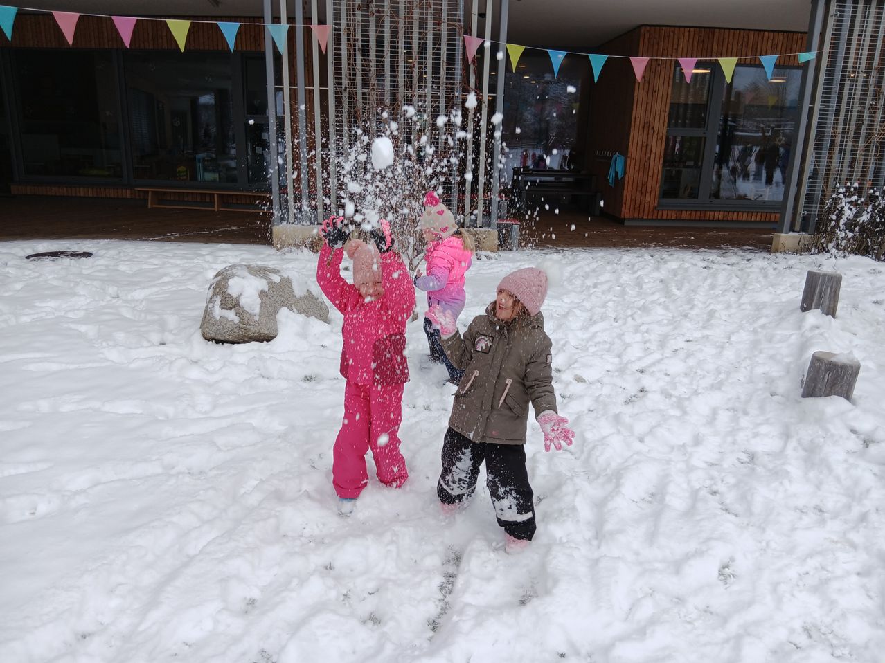 Drei junge Kinder in rosa Schneeanzügen werfen Schneebälle aufeinander. Sie sind draußen im Schnee in der Nähe eines Gebäudes.