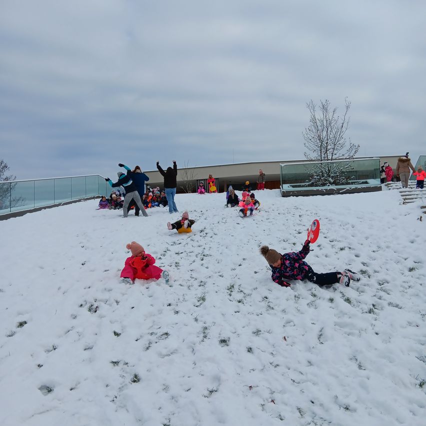 Eine Gruppe von Kindern spielt im Schnee auf einem Dach. Einige gleiten einen schneebedeckten Hang hinunter, während andere auf dem Boden liegen. Ein Erwachsener steht in der Nähe. Im Hintergrund befinden sich Glasbarrieren und ein Gebäude.
