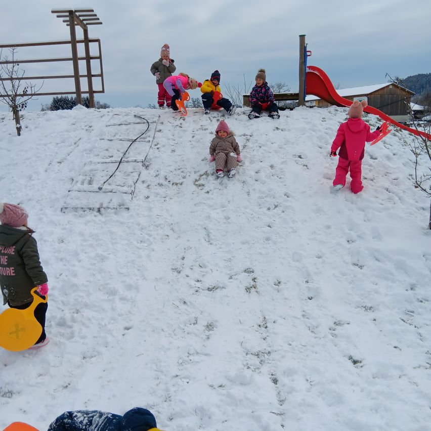 Kinder spielen im Schnee mit einer Rutsche im Hintergrund. Sie sind warm gekleidet in Mützen, Handschuhen und Stiefeln. Einige sitzen, während andere mit Schlitten stehen.