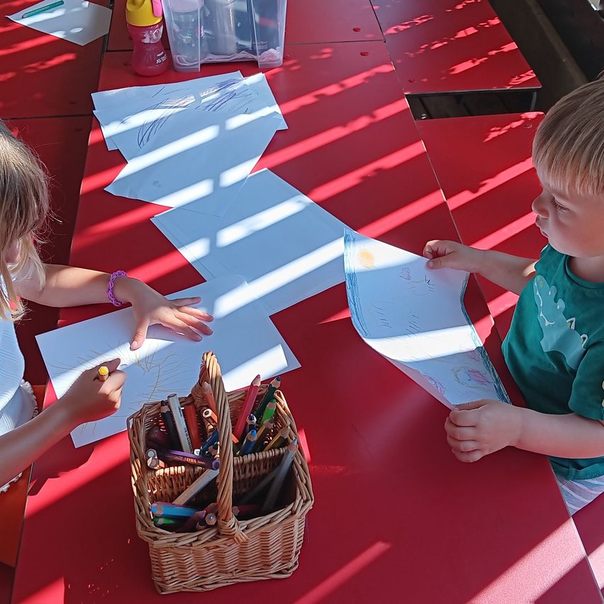 Two children are sitting at a red table, coloring in large sheets of paper. A basket of crayons is in front of them.