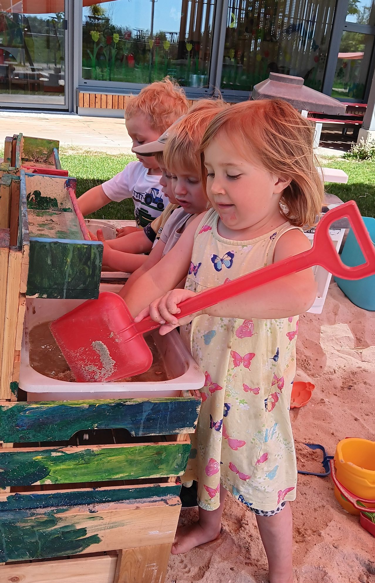 Three young children are playing in a sandbox, with a girl in a yellow dress holding a red shovel. Behind her, two boys are also playing in the sand.