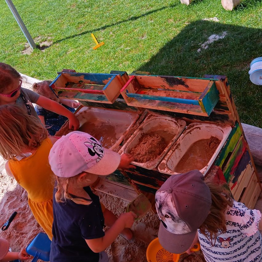 Several young children play in a sandbox with a wooden structure, wearing hats and holding shovels.