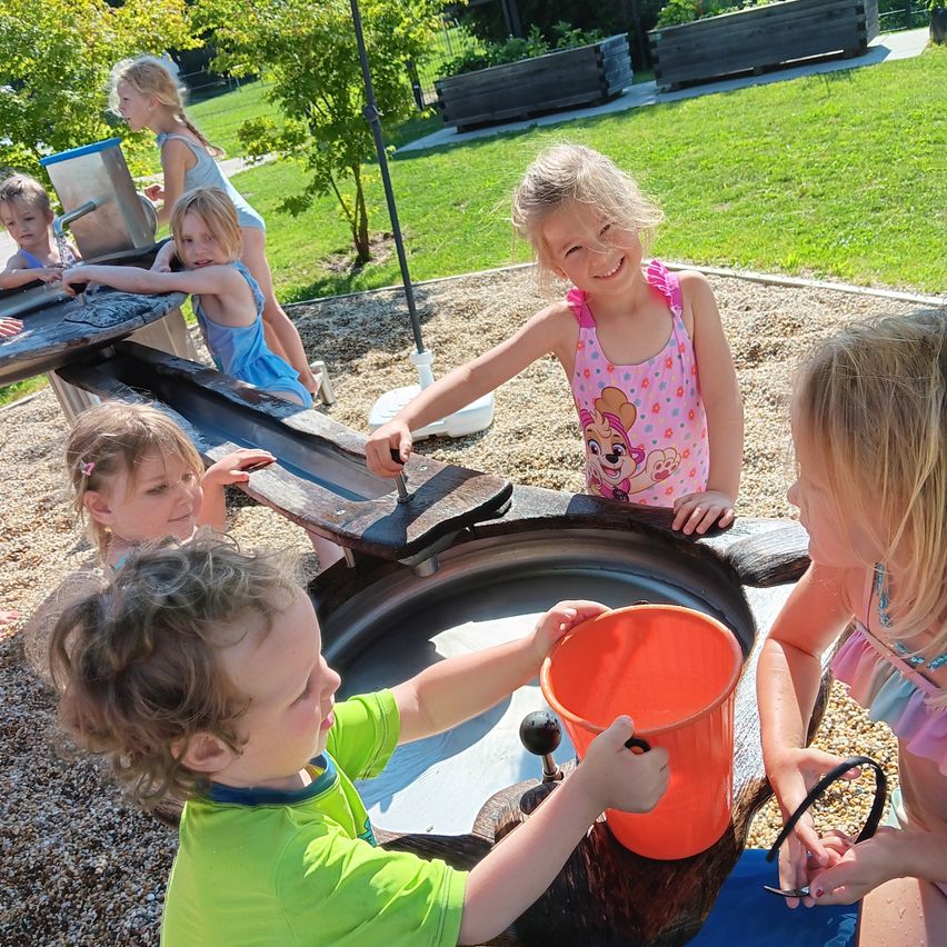 A group of young children play in a water play area, with one child using a bucket to pour water into a large basin.