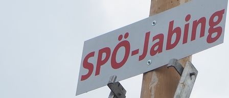 Ein Mann benutzt einen Bohrer, um ein Straßenschild für Spo-Jabing an einem Holzpfahl zu installieren, unter einem bewölkten Himmel.