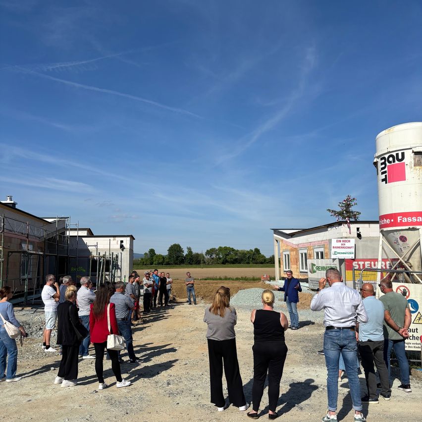 Eine Gruppe von Menschen ist unter blauem Himmel im Freien versammelt. Ein Mann spricht vor einer Baustelle mit einem großen weißen Silo zu ihnen.