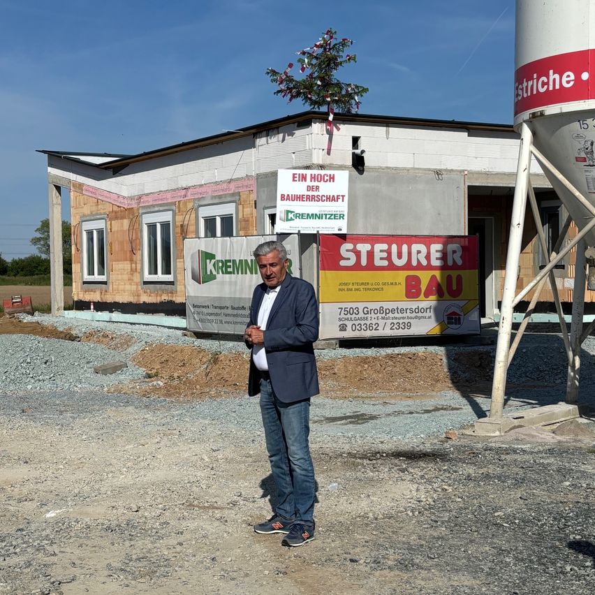 Ein Mann steht vor einem im Bau befindlichen Gebäude mit einem Schild 'Ein Hoch der Bauherrschaft' und einem Banner für 'Steuer Bau'. Das Gebäude hat einen Baum auf dem Dach und einen Silo.