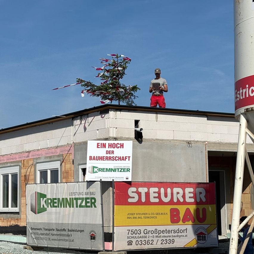 Ein Mann steht auf dem Dach eines Gebäudes im Bau. Das Gebäude hat mehrere Banner, darunter eines mit der Aufschrift 'Steuer Bau'. Es gibt zwei große Container, einer mit einem Schild für 'Kremmitzer' und ein anderer mit 'Ein Hoch der Bauherrschaft'.