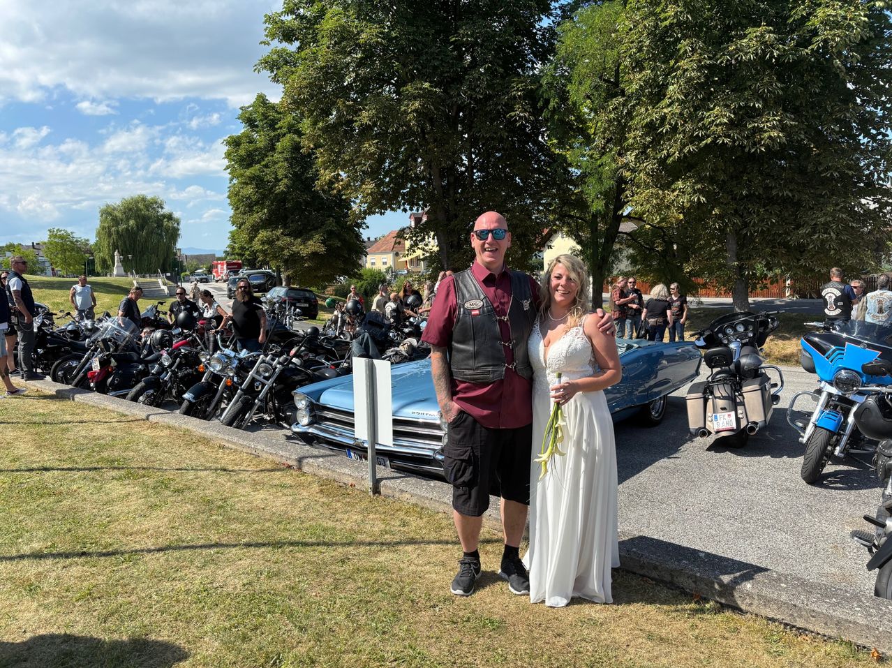 Ein Mann und eine Frau posieren für ein Foto vor einem geparkten blauen Auto und mehreren Motorrädern, mit Menschen, die dahinter stehen. Bäume, Gebäude und ein bewölkter Himmel sind im Hintergrund zu sehen.
