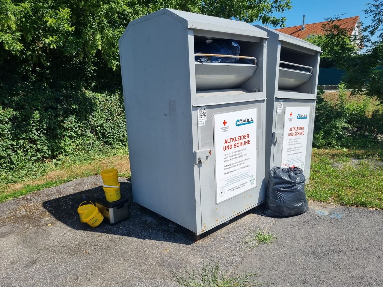 Zwei graue Boxen für das Recycling von alten Kleidungsstücken und Schuhen stehen im Freien. Ein gelber Eimer ist links. Ein schwarzer Müllsack ist rechts.