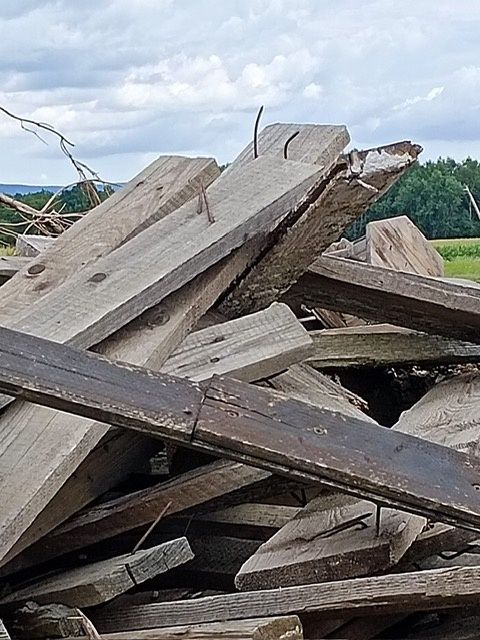 Ein Haufen alter, verwitterter Holzplanken ist zu sehen. Das Holz zeigt Anzeichen von Abnutzung und Alter, wobei einige Stücke zerbrochen sind. Der Ort scheint sich im Freien zu befinden, mit einem bewölkten Himmel im Hintergrund.