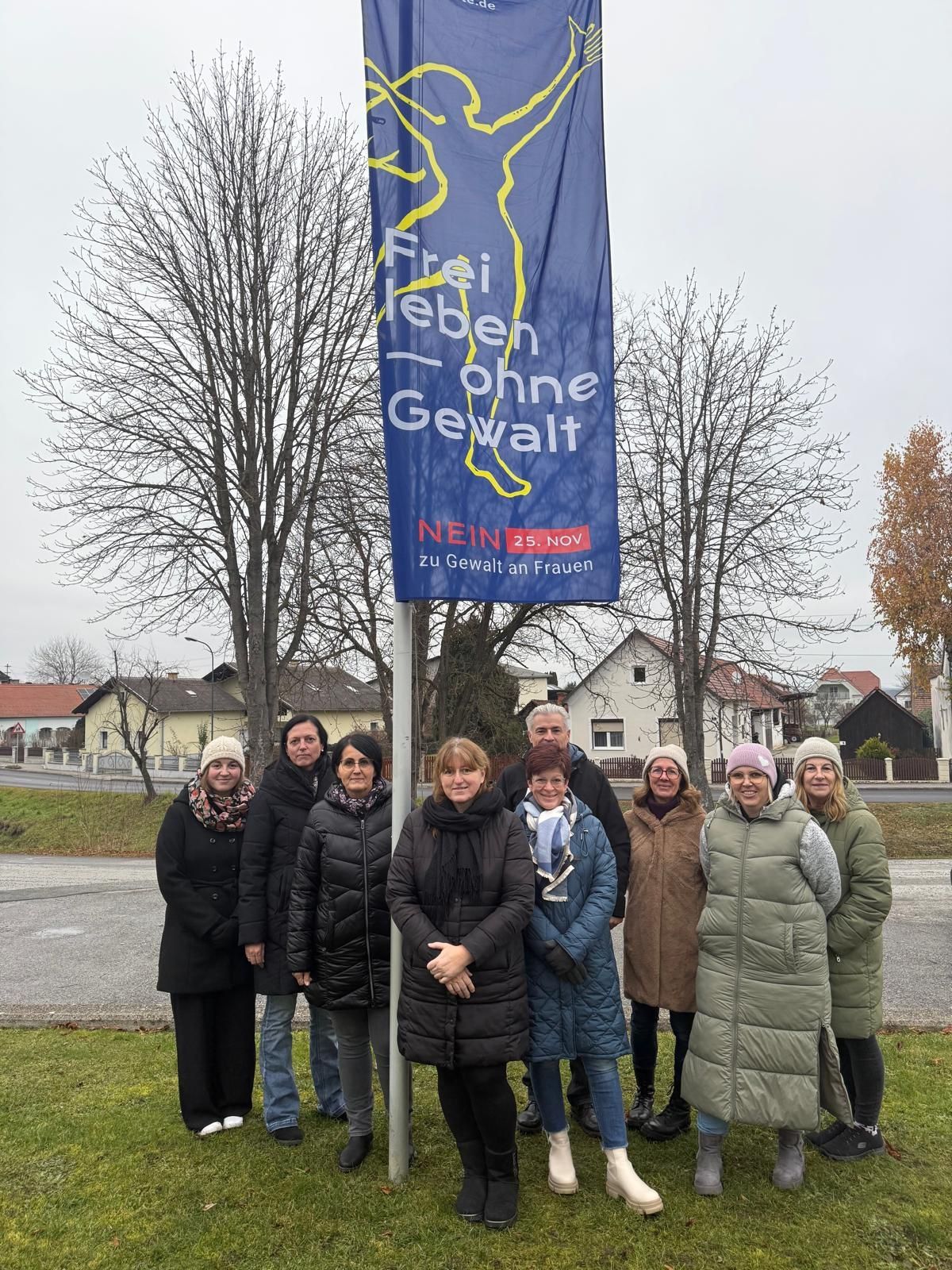 Eine Gruppe von Menschen steht bei einem Banner in einer Außenumgebung, mit dem Slogan 'Frei Leben Ohne Gewalt'. Sie sind warm gekleidet, mit Winterkleidung und Accessoires. Das Banner ist blau und trägt das Wort 'Nein', zusammen mit dem Datum '25. Nov'. Bäume und Häuser sind im Hintergrund zu sehen.