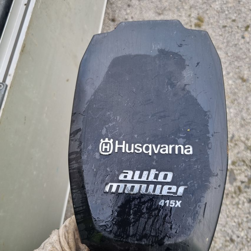 Ein schwarzes Objekt mit dem Husqvarna-Logo und dem Text 'auto mower 415X' wird auf einem strukturierten Hintergrund angezeigt.