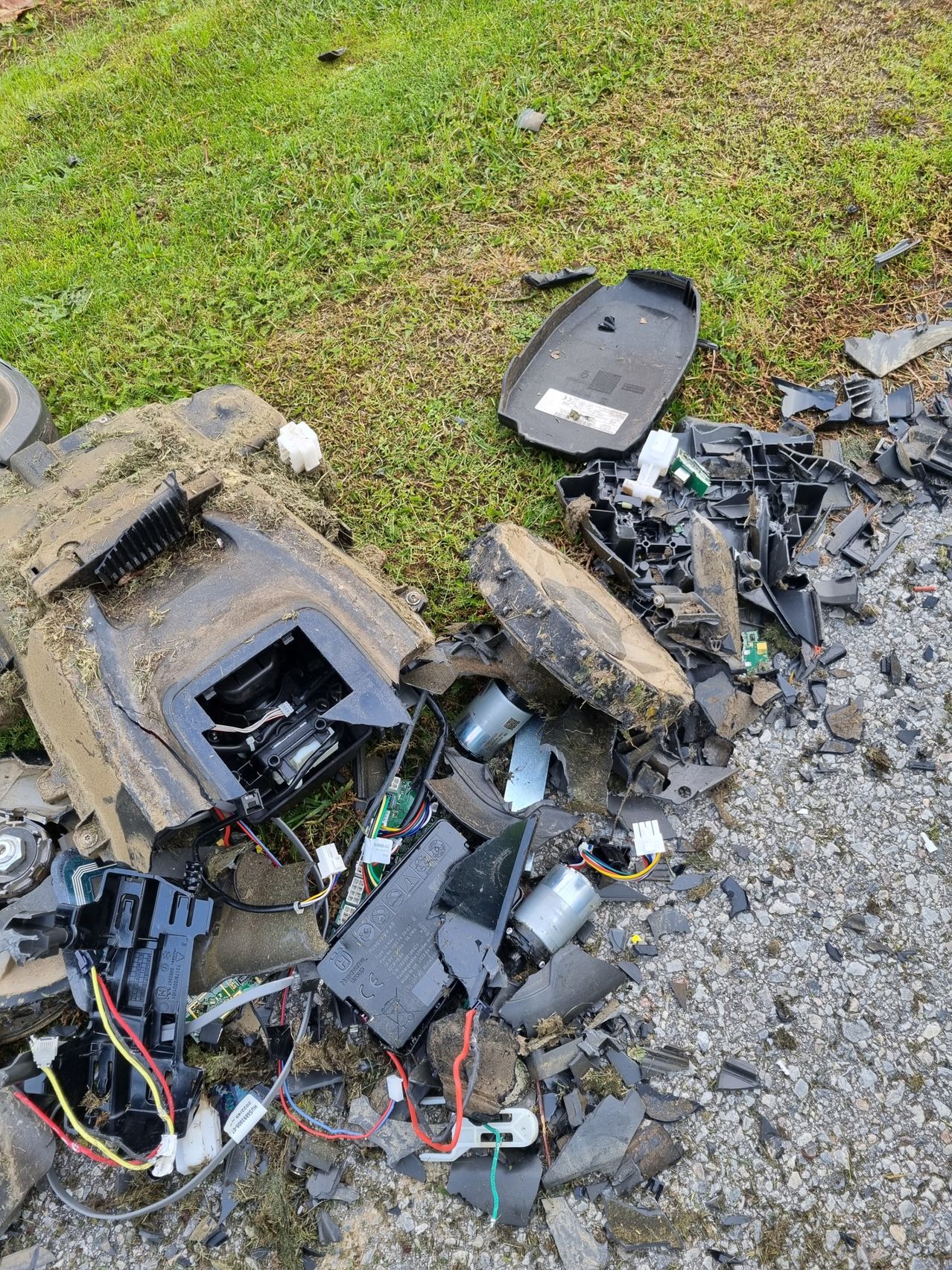 Verschiedene zerbrochene elektronische Komponenten und Teile, die auf dem Boden verstreut sind und mit Schmutz bedeckt sind. Die Stücke umfassen ein zerbrochenes Plastikcover, Leiterplatten, Kabel und andere elektronische Komponenten.