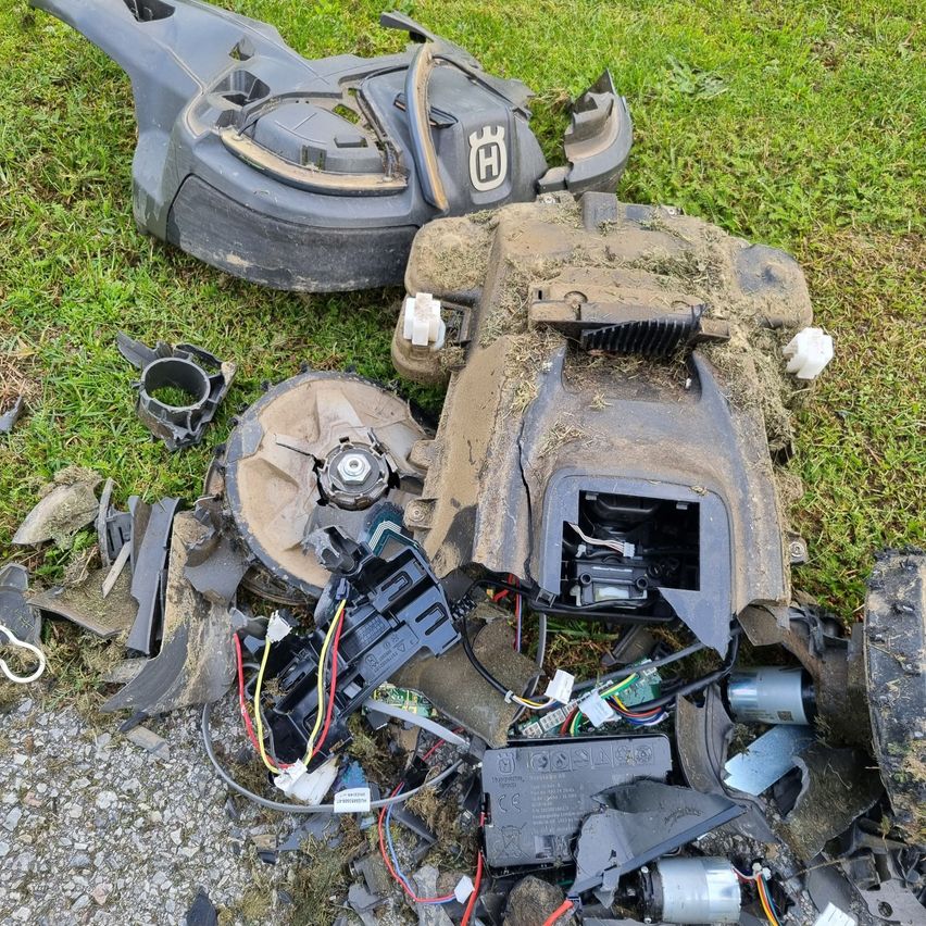 Mehrere Teile eines kaputten Rasenmähers sind auf einer grasigen Oberfläche verstreut. Das Kunststoffgehäuse und die Messer des Mähers sind zerbrochen, und die internen Komponenten sind freigelegt.