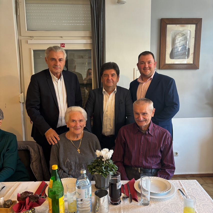 Eine Gruppe von Männern und eine ältere Frau posieren für ein Foto im Esszimmer. Der Tisch ist mit Tellern, Gläsern und einer Vase mit Blumen gedeckt. Ein Bilderrahmen ist an der Wand hinter ihnen montiert.