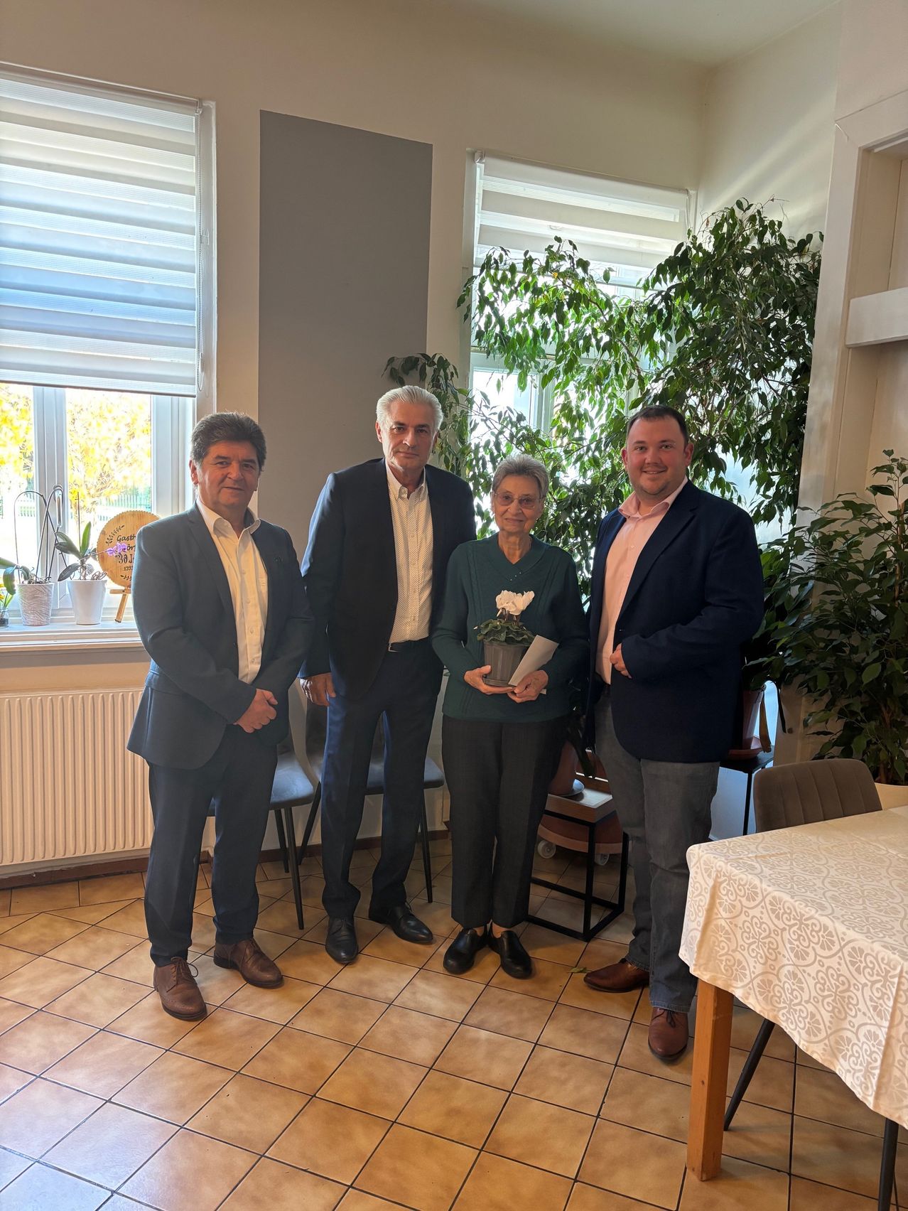 Vier Menschen stehen lächelnd für ein Foto in einem Raum. Eine Frau hält eine Topfpflanze, und sie stehen vor einem Fenster mit Pflanzen und einem Heizkörper. Ein Tisch mit einer weißen Tischdecke befindet sich rechts.