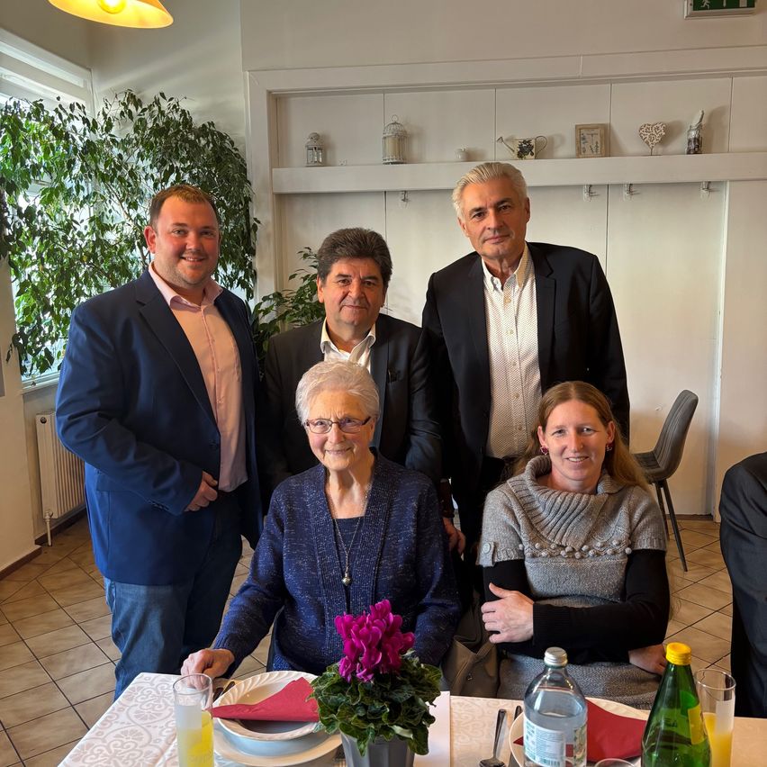 Fünf Personen sitzen und stehen um einen Tisch mit Blumen und Tellern herum. Zwei Männer stehen hinter einer älteren Frau. Eine weitere Frau sitzt neben ihr. Sie alle scheinen für ein Foto zu posieren. Dahinter befindet sich ein Regal mit Dekorationsartikeln.