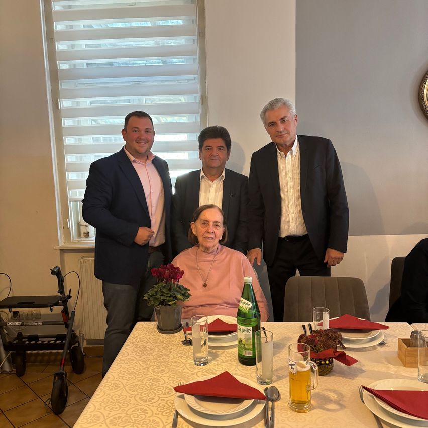 Vier Männer posieren in einem Raum mit einer lächelnden älteren Frau, die in einem Stuhl sitzt. Im Vordergrund steht ein Tisch mit Tellern, Gläsern und Blumen.