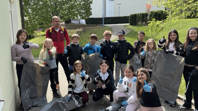 A group of children and adults hold plastic bags, wearing gloves. They stand on a sidewalk, with trees and buildings in the background.