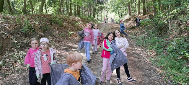 A group of children walks on a dirt path in a forest. Some wear gloves and carry plastic bags. The path is surrounded by greenery.