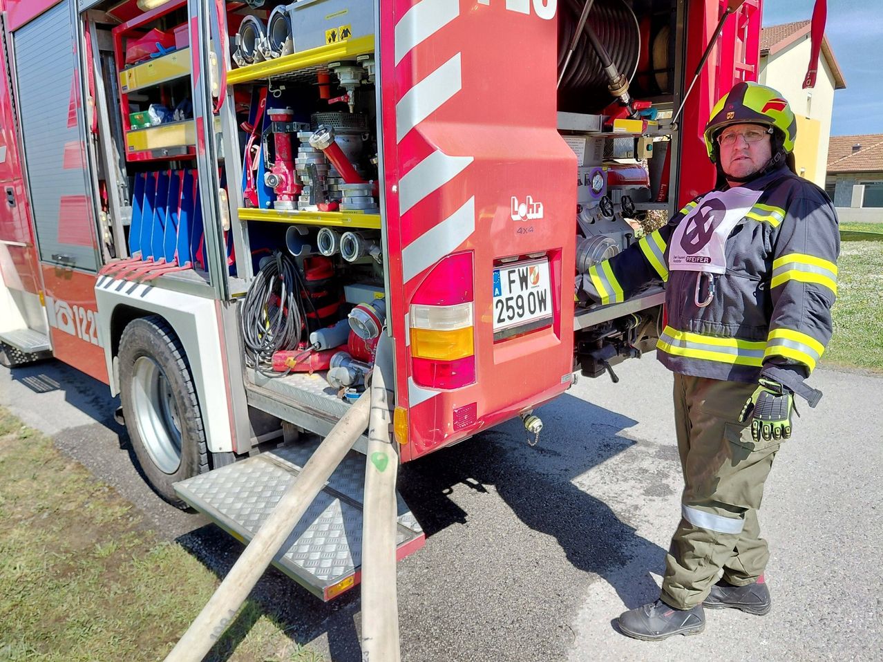 A firefighter stands next to a fire truck with its back door open, showing a variety of equipment. The license plate reads FW 259 OW.