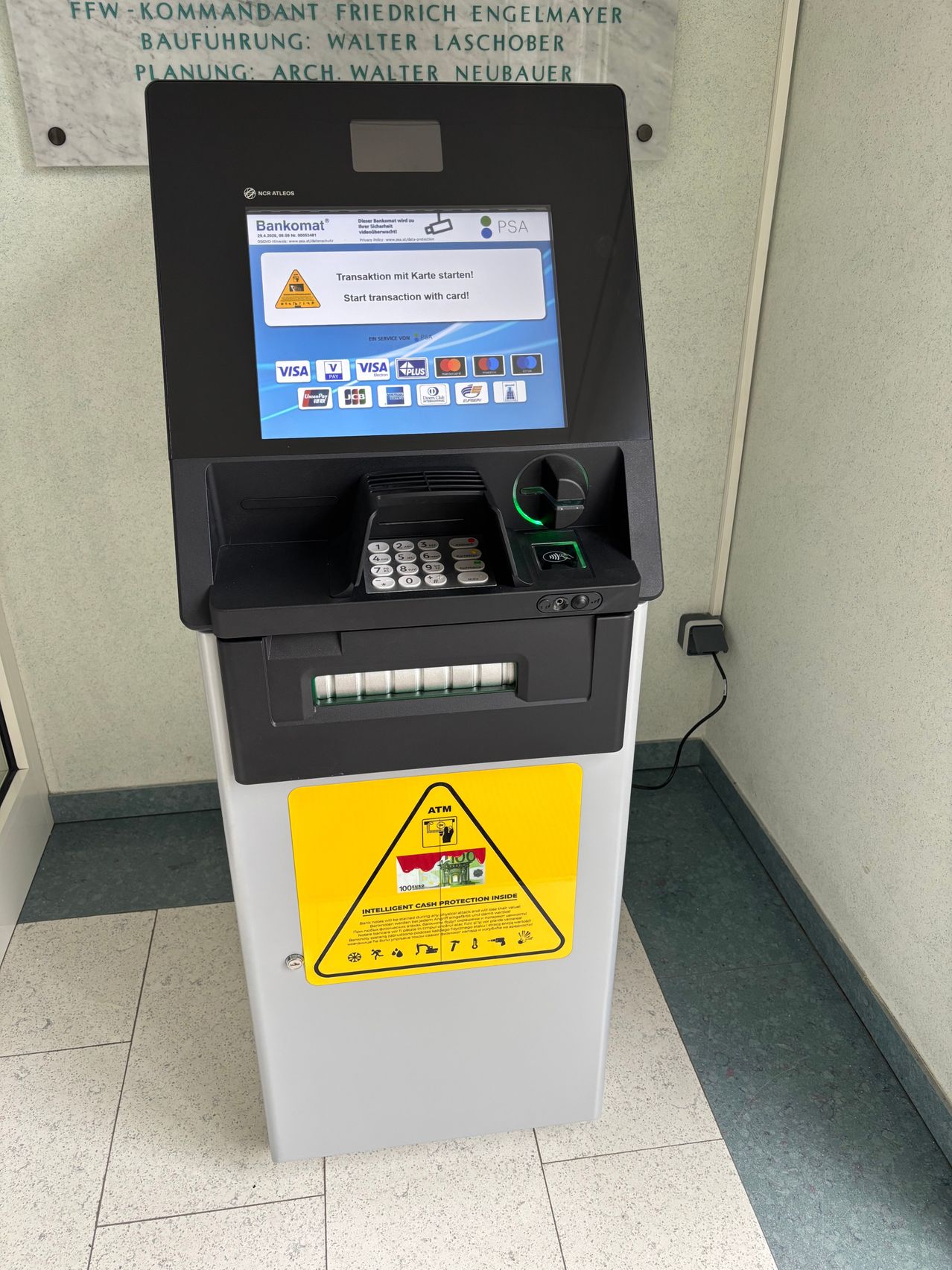 An ATM with a Bankomat screen displays a transaction start message. The machine has a keypad, card reader, and buttons for various functions. A yellow warning sign is attached to the front.