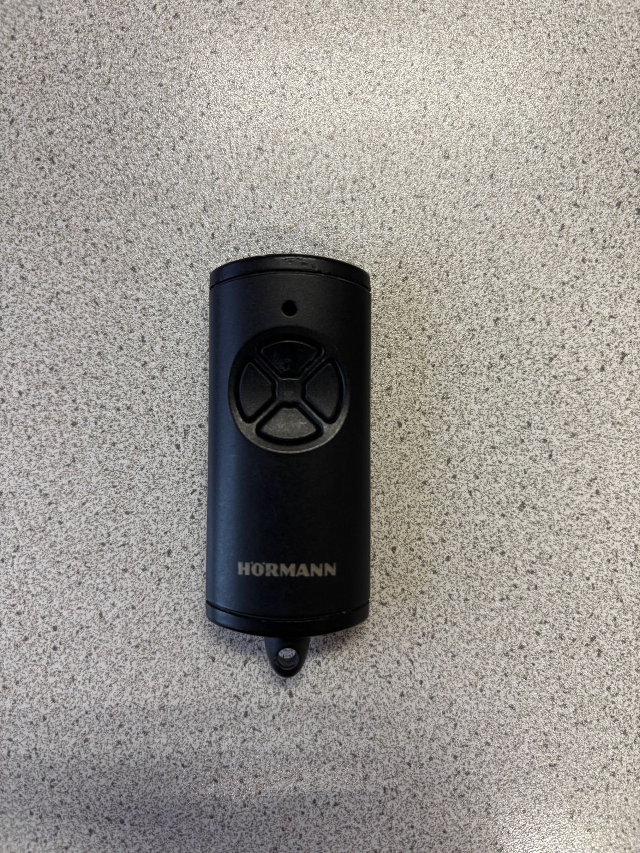 Eine schwarze Hormann-Fernbedienung liegt auf einer Marmoroberfläche. Sie hat einen runden Knopf und den Markennamen Hormann.