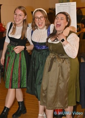 Drei Frauen in traditioneller Kleidung, mit Brille und Stiefeln, lachen zusammen in einem Raum mit Holzboden. Ein weißes Papier ist hinter ihnen, und ein Wasserzeichen lautet 'WUWU Video'.