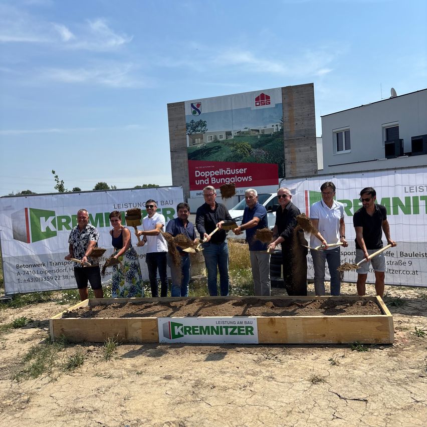 Eine Gruppe von Menschen in formeller Kleidung hält Schaufeln und steht um eine mit Erde gefüllte Holzkiste herum, möglicherweise für eine Grundsteinlegungszeremonie. Hinter ihnen befinden sich Banner und ein Gebäude mit einer großen Werbung.