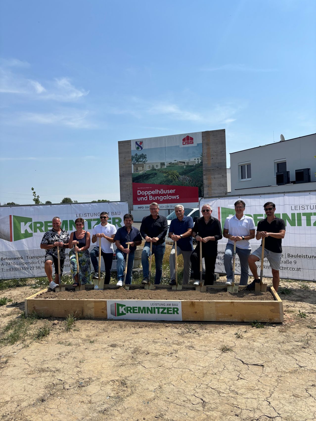 Eine Gruppe von Menschen in formeller Kleidung mit Schaufeln steht in einer mit Erde gefüllten Holzkiste, möglicherweise für eine Grundsteinlegungszeremonie. Hinter ihnen sind Banner mit den Texten 'Doppelhauser und Bungalows' und 'Leistung am Bau'.
