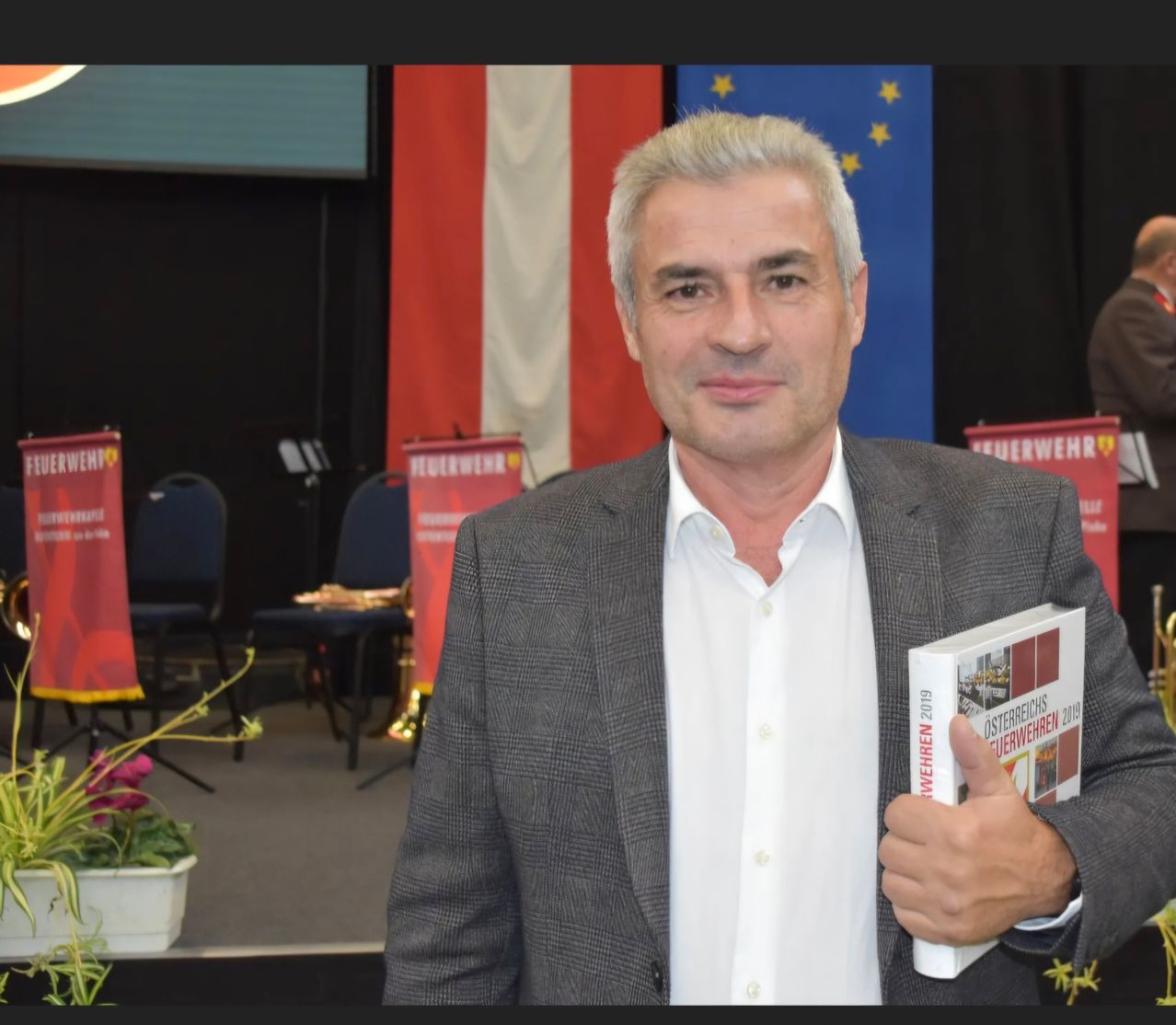 A man in a gray suit holding a book titled 'Feuerwehren 2019' is standing in front of a stage with a red and white backdrop.