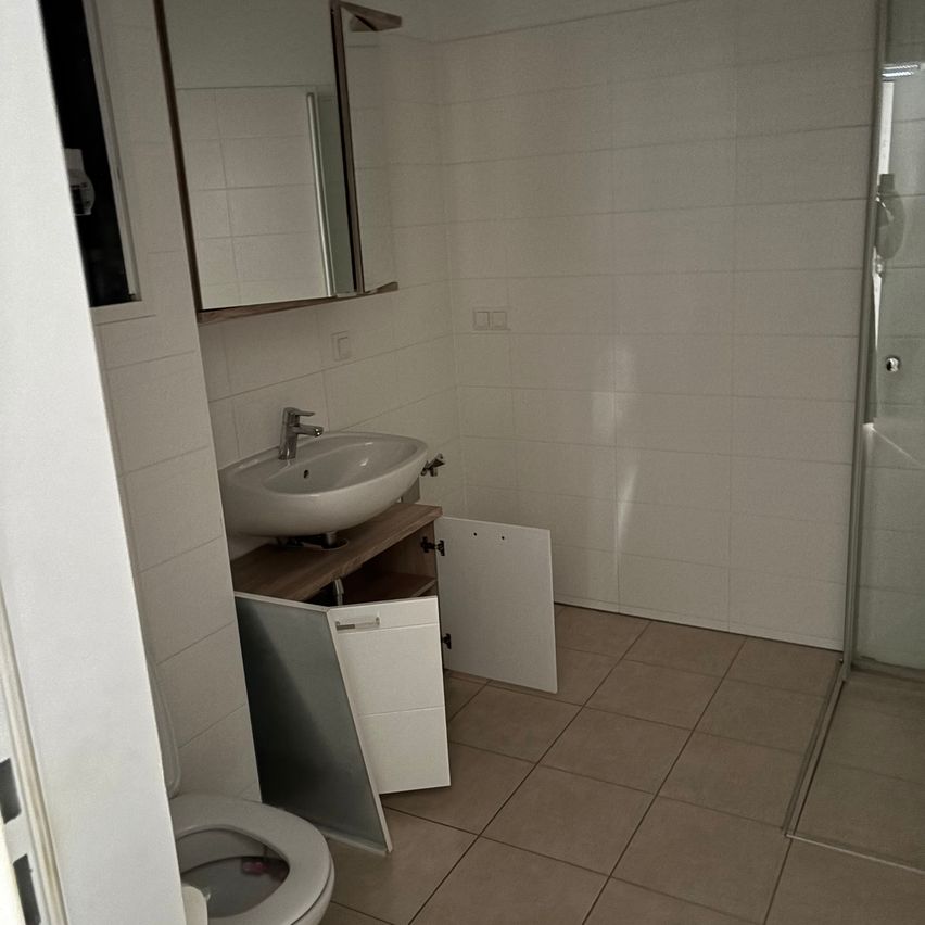 A small bathroom with white tiles, a sink, and an open cabinet below the sink. A toilet is partially visible on the left.