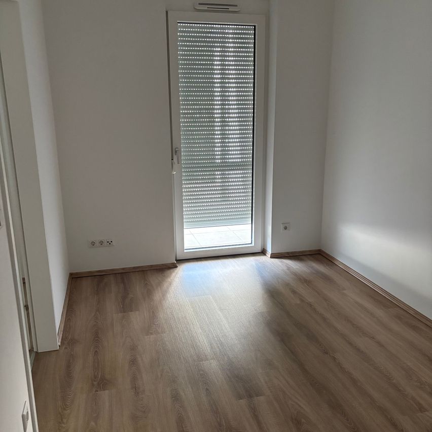 An empty room with white walls, wooden floors, and a glass door with blinds. The room has two power outlets on the left wall.