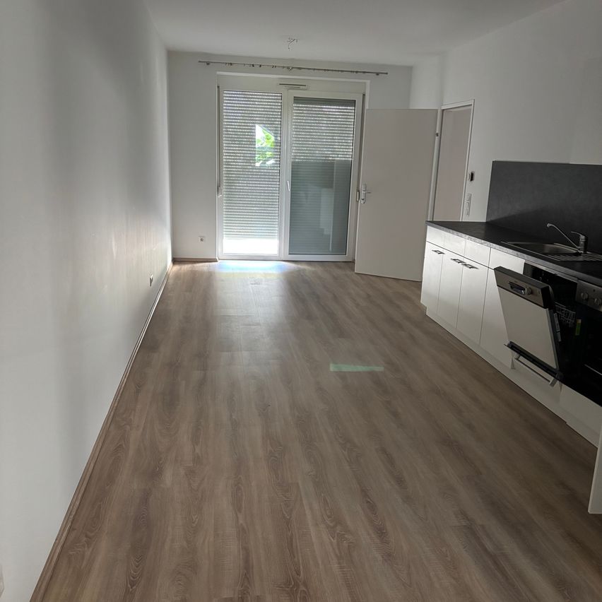 An empty kitchen with wooden floors, white walls, and a glass door leading to a balcony. The kitchen has a modern design with white cabinets and a black countertop.