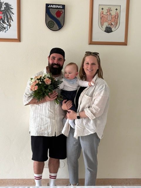 Ein Mann, eine Frau und ein Baby stehen vor einer Wand mit gerahmten Bildern. Der Mann hält Blumen und ein Baby, während die Frau ein Baby hält.