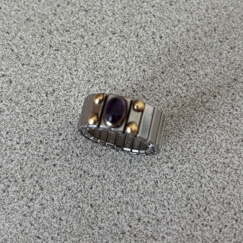 Ein silberner Ring mit goldenen Akzenten und einem violetten Stein in der Mitte liegt auf einer grauen Oberfläche.