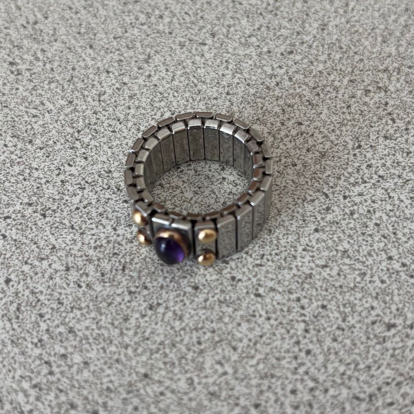 Ein Ring aus Silber und Gold mit einem lila Stein ist auf einer Marmoroberfläche ausgestellt. Der Ring besteht aus einer Reihe von miteinander verbundenen Metallstäben.