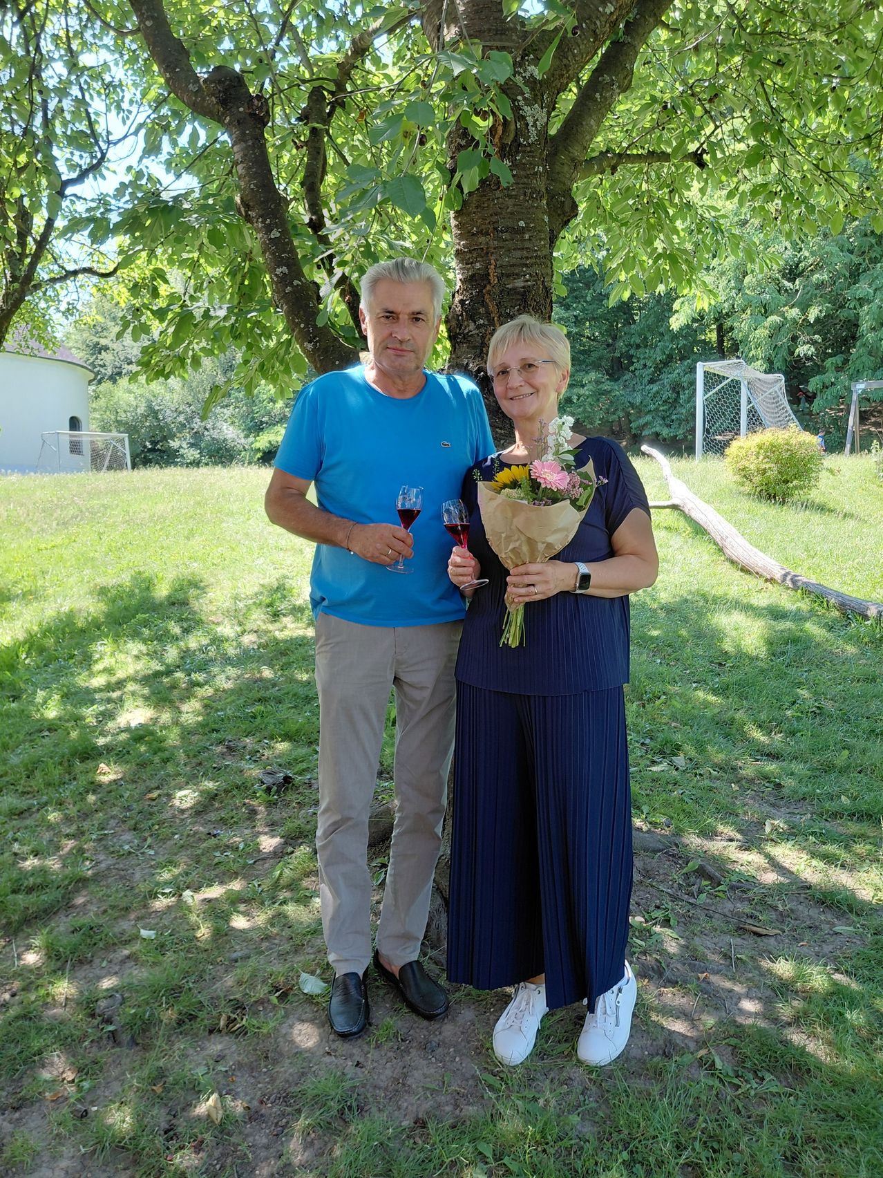 Ein Mann und eine Frau stehen lächelnd unter einem Baum. Der Mann hält Weingläser, die Frau einen Blumenstrauß. Sie tragen Freizeitkleidung. Ein Fußballtor und Bäume sind im Hintergrund.