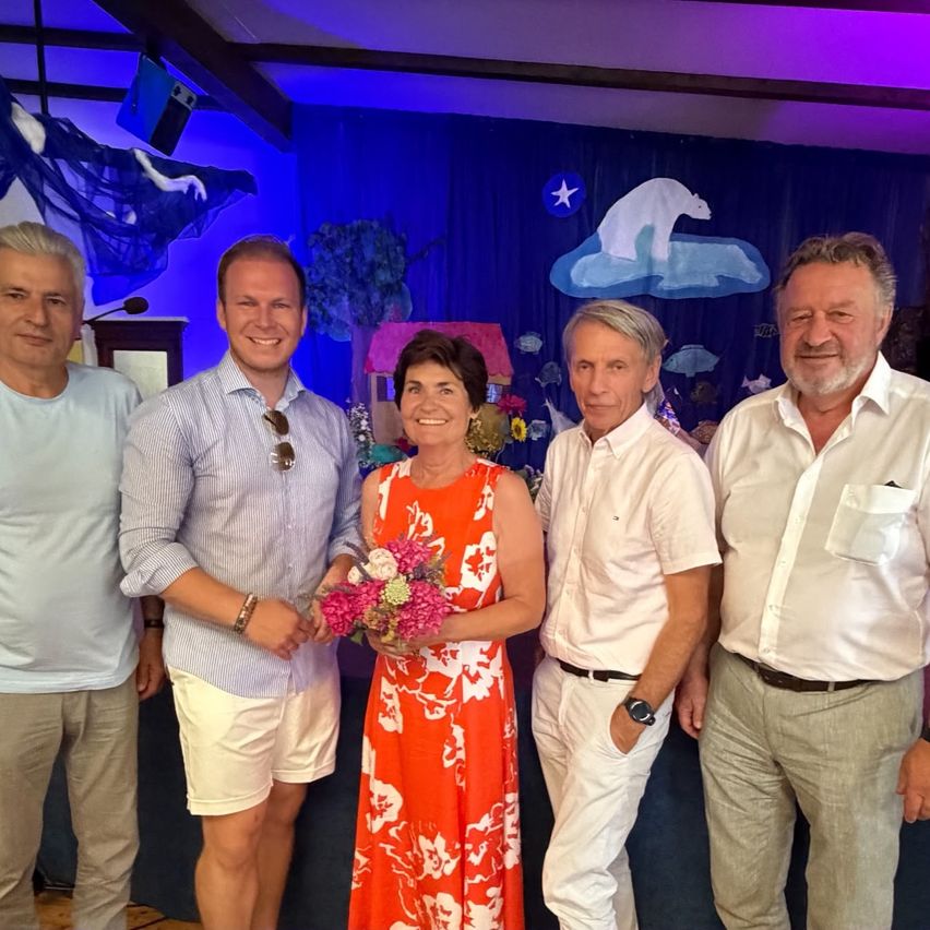 Fünf Erwachsene, zwei Männer und drei Frauen, stehen zusammen unter blauer Beleuchtung. Die Frau in der Mitte hält einen Blumenstrauß. Hinter ihnen ist eine Kulisse mit Zeichnungen eines Eisbären und eines Hauses.
